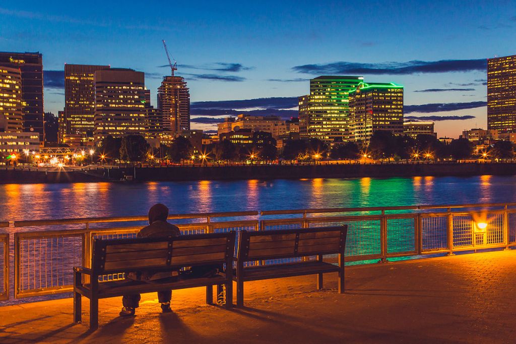 Evening cityscape of Portland from the Eastbank Esplanade with a person sitting on a bench.